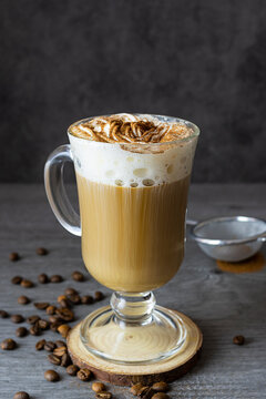 A High Glass Of Coffee And Slugs On A Wooden Stand. Coffee Beans On The Table. Strainer With Cinnamon In The Background. Dark Background