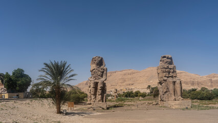 Giant sculptures of the colossi of Memnon. Huge dilapidated statues of seated pharaohs stand in the valley against the background of sand dunes and blue sky. Egypt