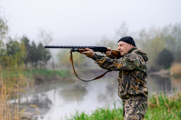 A male hunter with a rifle hunts and aims against the background of reeds and foggy lake