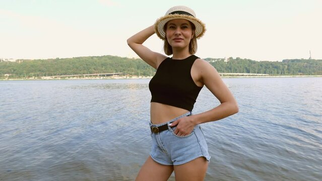 A young woman in short shorts and a black t-shirt in a hat stands on the beach near the river in the city. Slow motion. Relax in nature
