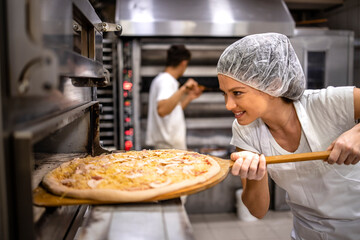 Working in pizza restaurant. Female chef in white uniform and hairnet putting pizza in the oven for...