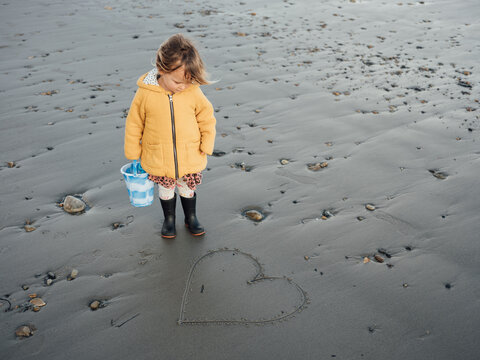 Young girl stands on a beach looking at a heart drawn in the sand.