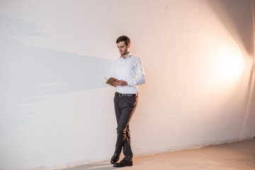 A man standing by the white wall, reading a book