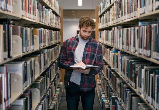 Male Student Reading Book In Library Stacks