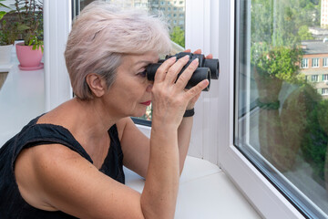 Portrait of an elderly woman 60-65 years old looking through binoculars from the window of her apartment. Concept: spying on neighbors, gathering information, espionage and gossip. - obrazy, fototapety, plakaty