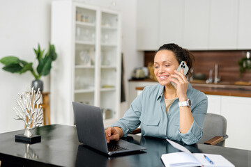 Obraz premium Side down shot of a woman talking on the phone while working on the laptop. She sits at the wooden table and smiling while typing at the laptop with her hand