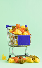 Colored vegetable pasta, in a small shopping cart and scattered on the green background.