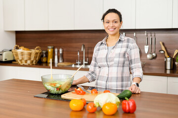 Woman is standing near the bowl of super healthy salad and smiling, she is in casual clothes in a cozy kitchen. Healthy eating concept