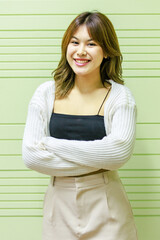 Portrait closeup studio shot of Asian young beautiful cheerful female model in black camisole shirt, white sweater and shorts standing smiling look at camera in front striped green wall background