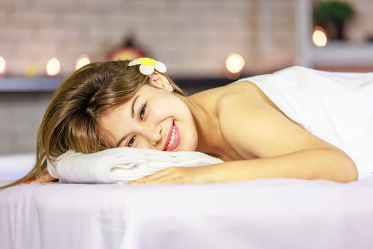 Closeup Asian young beautiful sexy happy female spa customer covered by white clean bath towel laying down resting relaxing closed eyes smiling on massaging bed putting bloom orchid flower on ear