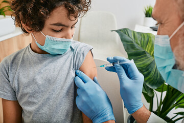 Routine vaccinations for a child. Teenage boy wearing medical mask while vaccinated by mature...