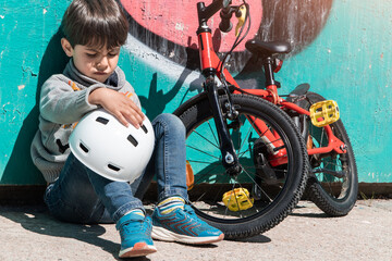 sad and angry little boy with bycicle and helmet sitted on the ground of the city. Child in a...