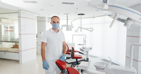 A young dentist in a mask stands near a red dental chair and smiles in modern white dentistry. Modern dentistry and prosthetics.