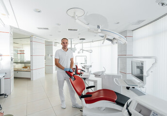 A young dentist stands near a red dental chair and smiles in modern white dentistry. Treatment and...