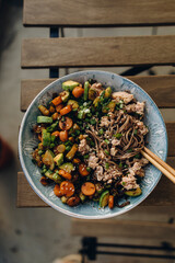 wok cooked vegetables and soba in a plate on a wooden table. hold soba with wooden chopsticks. soba with tuna and vegetables.