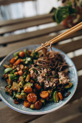 wok cooked vegetables and soba in a plate on a wooden table. hold soba with wooden chopsticks. soba with tuna and vegetables.