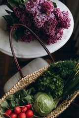 straw bag full of farm organic vegetables and herbs. a bunch of radishes, young cabbage and greens in a bag top view. kitchen table with a bouquet of lilacs and a bag of seasonal products