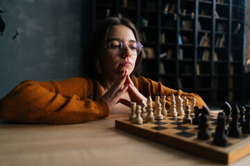 Thoughtful cute young woman in elegant eyeglasses thinking about chess move sitting on floor in...