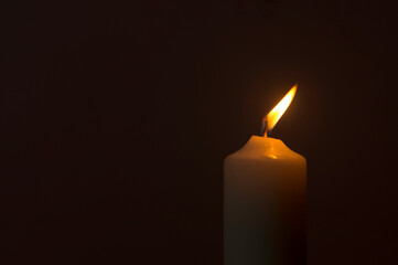 A single burning candle flame or light glowing on a white candle on black or dark background on table in church for Christmas, funeral or memorial service