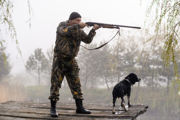 Hunter with a gun aims at the bridge with his dog Russian Spaniel on the background of sunrise and...