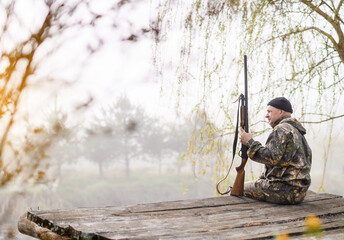 Male hunter sitting on a bridge against the backdrop of sunrise and forest with a lake. High...
