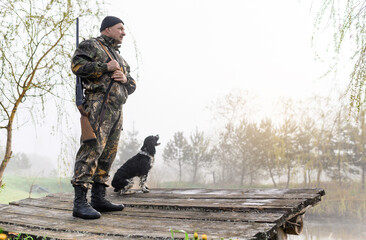 Hunter with a gun stands on the bridge with his dog Russian Spaniel on the background of sunrise...