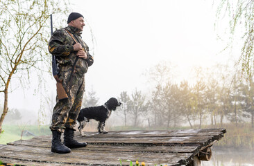 Hunter with a gun stands on the bridge with his dog Russian Spaniel on the background of sunrise...
