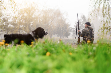 Male hunter sitting on a footbridge on a background of sunrise and forest with a lake in the foreground blurred dog spaniel