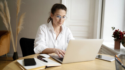 A woman with glasses is a freelancer working in an office using a laptop, making a project report - obrazy, fototapety, plakaty