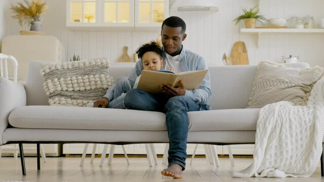 Mixed race boy sitting in arms of loving African American father and reading book together while sitting on bed at home. Black skin father with mixed race son reading book while sitting on couch.