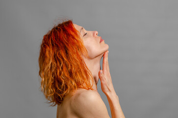 Naked redhead woman touches her chin. Sexy woman. Romantic look. Side view. Gray background.