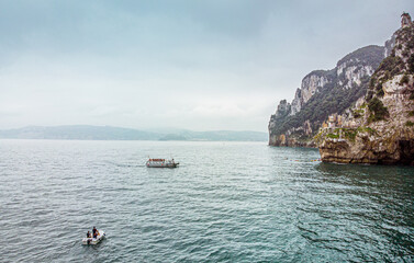 Small boat or yacht sailing alongside a ship on the sea near the cliffs where a lighthouse is located. Shot made with an aerial drone. Adventure of a cloudy day with waves.