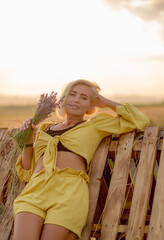 Pretty young woman in yellow among rural field with golden oat field on sunset background