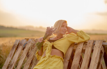 Pretty young woman in yellow among rural field with golden oat field on sunset background