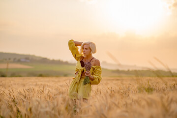 Pretty young woman in yellow among rural field with golden oat field on sunset background