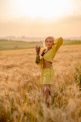 Pretty young woman in yellow among rural field with golden oat field on sunset background