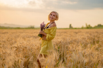 Pretty young woman in yellow among rural field with golden oat field on sunset background