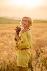 Pretty young woman in yellow among rural field with golden oat field on sunset background