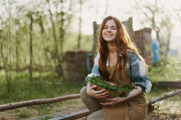 A young woman feeds her chickens on the farm with grass, wearing a simple plaid shirt, pants and apron, and smiling for the camera, caring for the animals