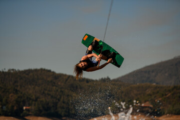 Active guy wakeboarder flips on wakeboard over splashing wave against sky and hills - obrazy, fototapety, plakaty