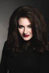 Fototapeta premium Beautiful brunette woman with long dark fluffy, wavy hair and green eyes studio portrait. Model with evening make-up and red lipstick wearing dark clothes in dark studio background looking to camera