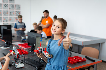 Portrait girl student holds a robot in her arms, which was assembled from plastic parts programmed on a computer in a robotics lesson and shows a thumb up