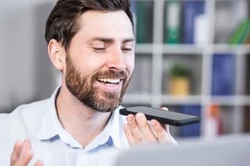 Cheerful and happy man records audio messages, freelance businessman in office, uses application on smartphone