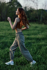 A woman with long red hair works out and runs on the green grass in the park in sweatpants and sneakers