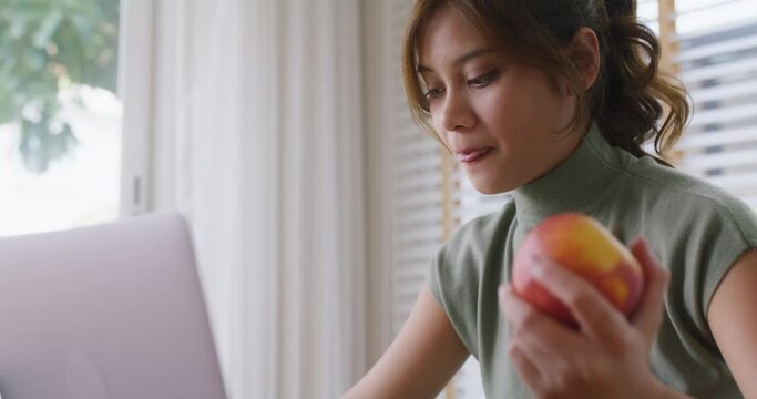 Close-up young cute girl asia people busy work sit at home table hold organic red apple on hand bite full mouth eat fresh fruit snack for weight loss low calories immune vitamin good fiber diet meal. 