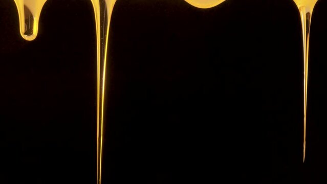 Drops of thick golden honey flowing down on a black background. Close up of sweet nectar or molasses being spilled. Liquid honey pours and drips. Sugar syrup is pouring. Healthy and curative dessert.