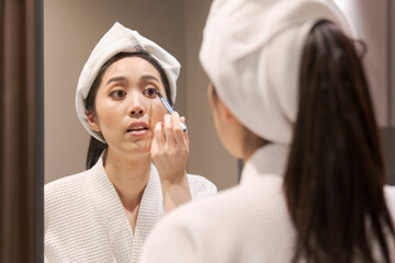 Beautiful young woman wrapped in a towel doing her makeup in the morning. Asian woman wrapped in white towel applying makeup after shower. Beautiful girl putting daily makeup.