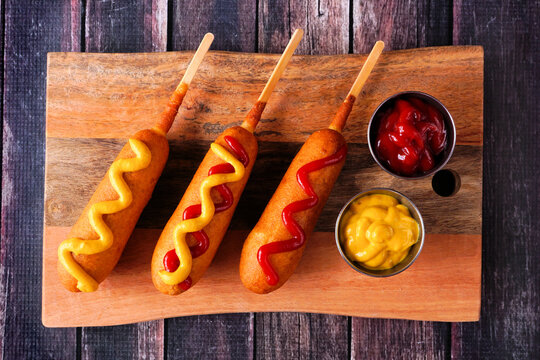 Corn dogs with different toppings on a wooden platter. Overhead view on a dark wood background.