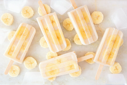 Healthy banana yogurt popsicles. Above view with scattered fruit slices and ice cubes over a bright white marble background.