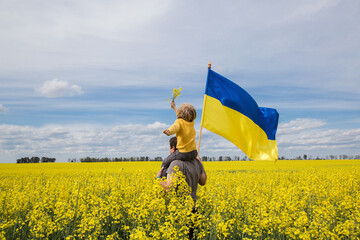 dad with his son sitting on his shoulders with a large satin flag of Ukraine in hands among a blooming yellow rapeseed field. Education of patriotism. Pride, freedom. Ukrainians against the war